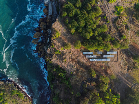 Aerial view of Baron Beach featuring a solar panel array on the cliffside near the ocean, surrounded by trees and rugged terrain, Yogyakarta, Special Region of Yogyakarta, Indonesia.