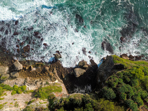 Aerial view of Baron Beach with dramatic rocky cliffs, lush green vegetation, and turquoise waves crashing against the shore, Yogyakarta, Special Region of Yog, Indonesia.