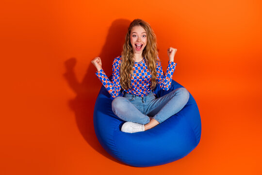 Excited young woman in a stylish pullover sitting on a beanbag against a vibrant orange background