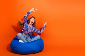 Fotobehang Sportwinkel Excited Young Woman in a Stylish Checkered Pullover Posing on a Bean Bag with Bright Orange Background  © deagreez