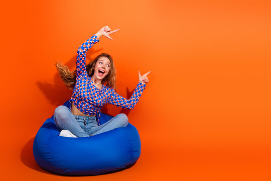 Excited Young Woman in a Stylish Checkered Pullover Posing on a Bean Bag with Bright Orange Background