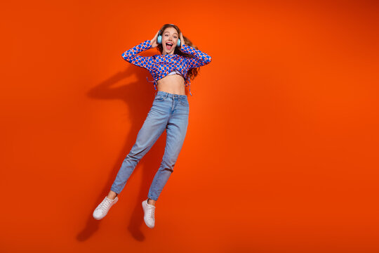 Young woman in casual checkered top and jeans enjoying music with headphones against vibrant orange background