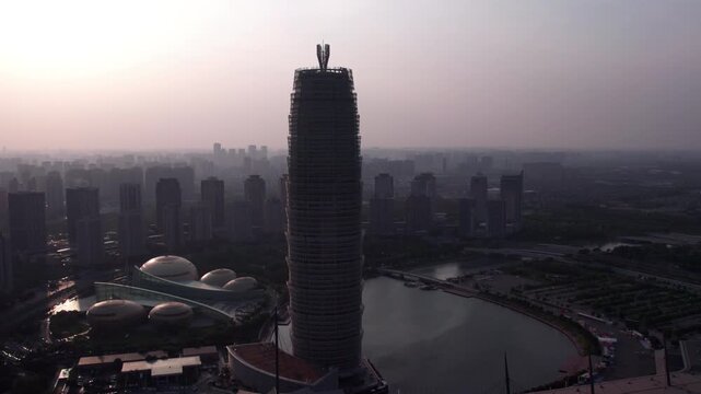 Aerial view of a modern urban district featuring a prominent tall skyscraper beside a water body, surrounded by city buildings under hazy dusk sky.
