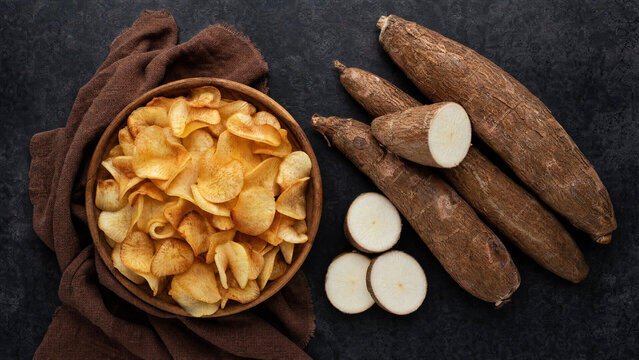 Crispy cassava chips in wooden bowl with whole and sliced raw yucca roots on dark background