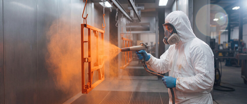 Powder coating metal frame in factory workshop by worker in white protective suit spraying bright orange paint using electrostatic spray gun for manufacturing and industrial finishing