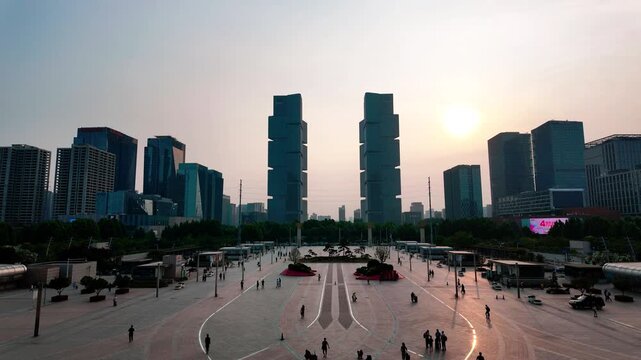 A wide urban public plaza flanked by modern skyscrapers, with pedestrians walking under soft morning or evening light.
