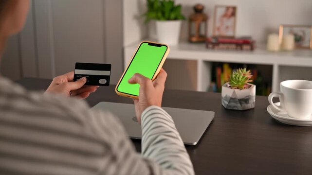 Woman holding smartphone with green screen and payment card near laptop, coffee cup and plant. Mobile payment scene supports online shopping concepts, digital checkout and secure commerce visuals