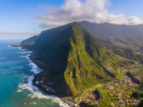 Aerial view of the dramatic green cliffs of Madeira plunging into the Atlantic Ocean with a coastal town nestled at the base under a bright blue sky in Madeira, Madeira, Portugal.