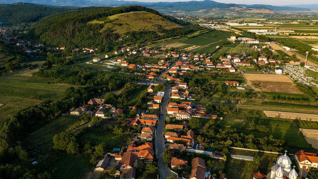 Aerial view of the town of Deva nestled in the rolling hills, featuring traditional houses with red-tiled roofs, surrounding agricultural fields, and a small church, Deva, Hunedoara, Romania.