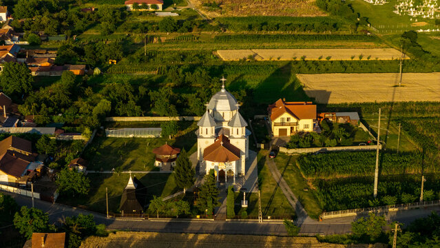 Aerial view of a traditional Orthodox church surrounded by lush green fields, residential houses, and trees in the golden hour light in Deva, Hunedoara, Romania.