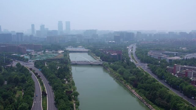 An aerial shot shows a river running through an urban area, flanked by roads, green spaces and distant city buildings under hazy sky.