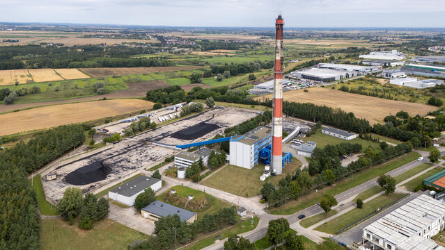 Aerial view of an industrial facility featuring a tall red-and-white chimney, large coal storage piles, and surrounding green fields and warehouses in Tczew, Poland.