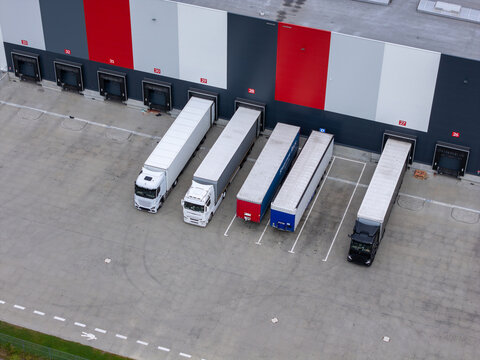 Aerial view of a modern logistics warehouse with semi-trucks parked at numbered loading docks on an asphalt yard in Tczew, Poland.