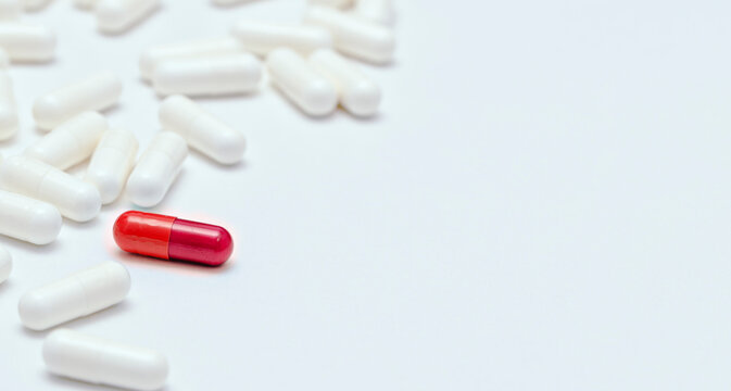 Close up of a single distinctive red pill capsule standing out among many white capsules on a white background, uniqueness and medical concept.