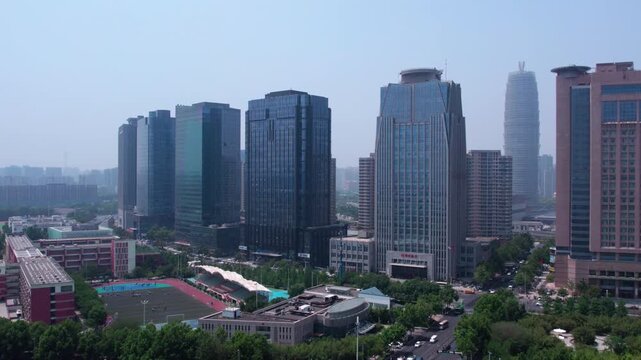 A daytime city view featuring modern glass skyscrapers, a busy road with traffic, and green trees along the street.