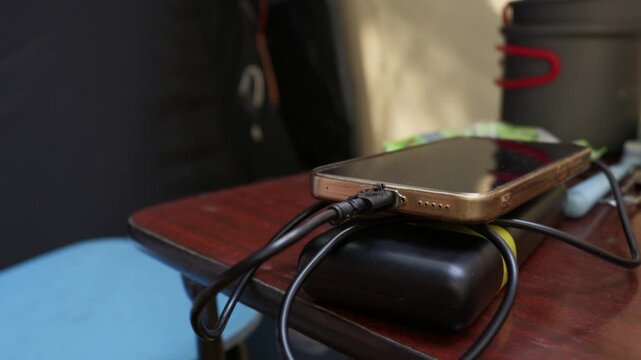 A smartphone charges from a portable power bank on a wooden table at a campsite with a fly sitting on the cable. Concept of off-grid energy, travel technology, and outdoor adventure.