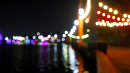 Blurred perspective view looking along a lively waterfront at night, with strings of colorful party lights and promenade lights creating soft bokeh effects and long reflections in the water. © gusense