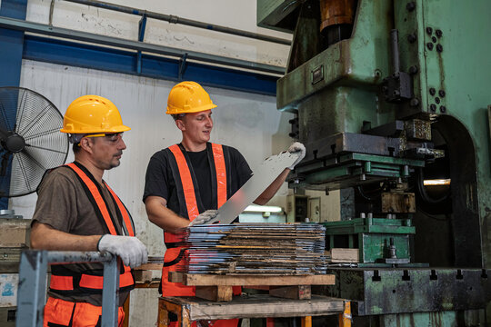 Two professional male industrial factory workers wearing yellow safety helmets carefully inspect a steel sheet metal part near a heavy automated stamping press machine during production.