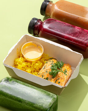 Healthy meal box with fish cutlets yellow turmeric rice sauce and assorted detox smoothie bottles on green background