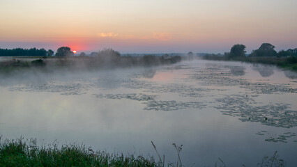 A serene river reflects the warm glow of a rising sun while mist gently drifts across the calm water at dawn © Александр Арендарь