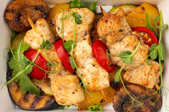 Healthy meal box with grilled chicken breast skewers roasted vegetables mushrooms and fresh greens close up
