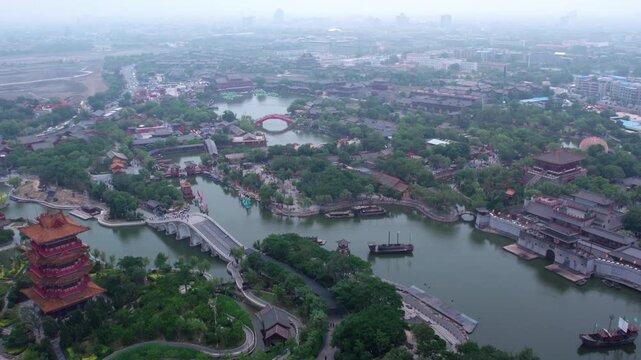 An aerial shot of a traditional water town featuring historic architecture, winding waterways, boats and lush greenery under a hazy sky.