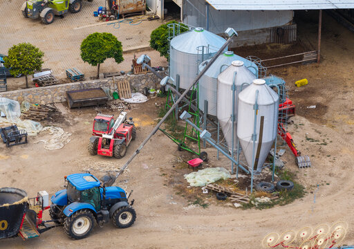 Aerial view of a farm with metal grain silos, a blue tractor, and a red telehandler on a dirt lot in Sopotnitsa, Municipality of Demir, North Macedonia.