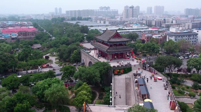 Aerial view of a traditional Chinese ancient gate tower, surrounded by green parks and modern urban buildings in the distance.