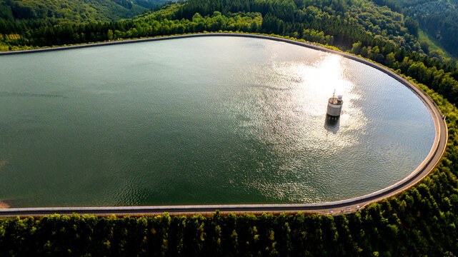 Aerial view of a pumped storage power plant reservoir surrounded by dense green forest with sunlight reflecting on the water in Germany.