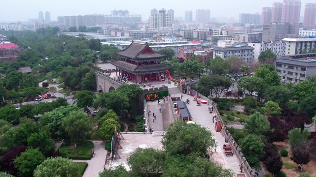 An aerial view of a traditional ancient gate tower standing in a green urban park, with city buildings in the distance.