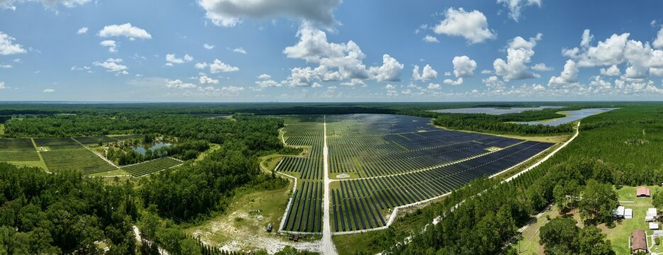 Aerial view of Magnolia Solar Farm with rows of photovoltaic panels set amidst lush green forests and open fields under a bright blue sky in Green Cove Springs, Florida, United States.