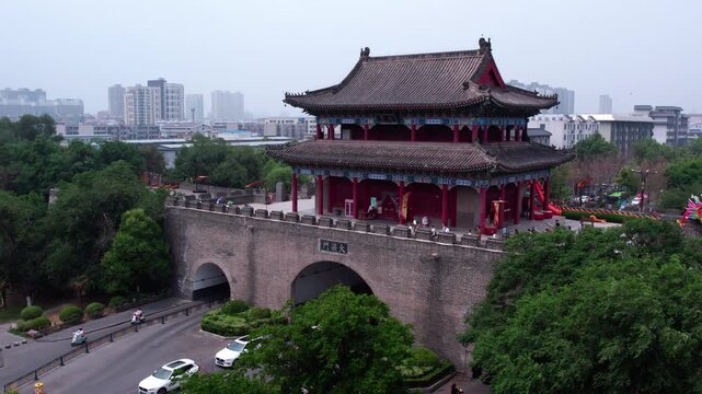 A traditional ancient city gate tower stands with a modern urban backdrop, surrounded by greenery and people visiting the landmark.