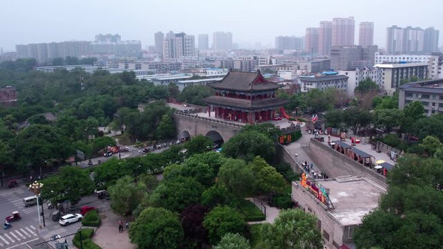 A high-angle view of a traditional Chinese city gate tower, set in a green urban area with surrounding modern buildings under a hazy sky.