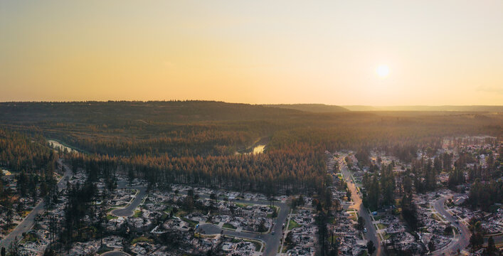 Aerial view of the aftermath of the Spokane wildfire, showing destroyed residential homes, scorched pine trees, and a river under a hazy sunset sky in Spokane, Washington, United States.