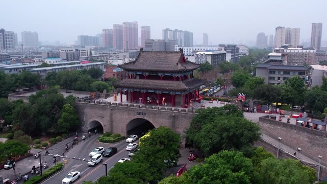 A historic city gate tower stands with modern urban buildings and traffic around it, surrounded by green trees.