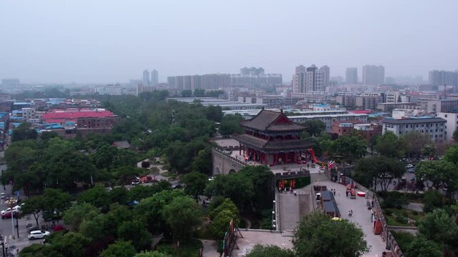 A historic traditional Chinese pavilion stands in a green urban park, with modern city buildings in the hazy background.