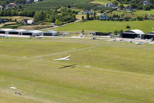 Aerial view of a glider flying over the grassy runway at Aeroklub Podhalanski im. Leopolda Kwiatkowskiego with hangars and small aircraft in Lososina Dolna, Malopolskie, Poland.