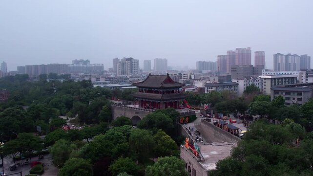 A traditional ancient Chinese tower stands amid dense urban greenery, with modern city buildings in the distance.