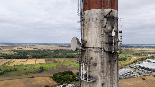 Aerial view of a weathered industrial chimney with communication antennas and ladders, overlooking the surrounding agricultural fields and rural landscape in Tczew, Poland.