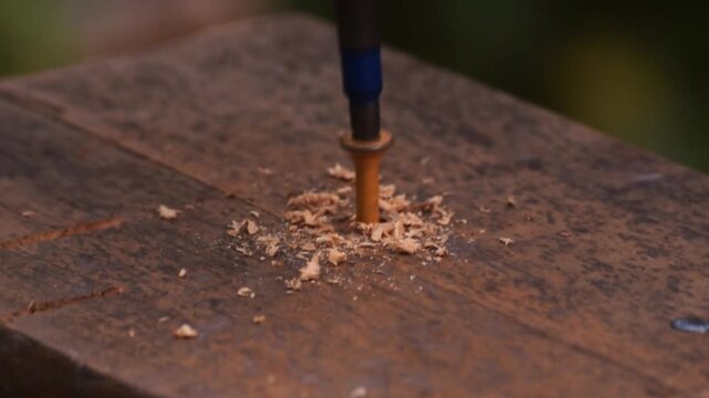 Close-up angled view of a power drill driving a screw into a wooden board, with wood shavings collecting around the screw as it penetrates the surface in slow motion.