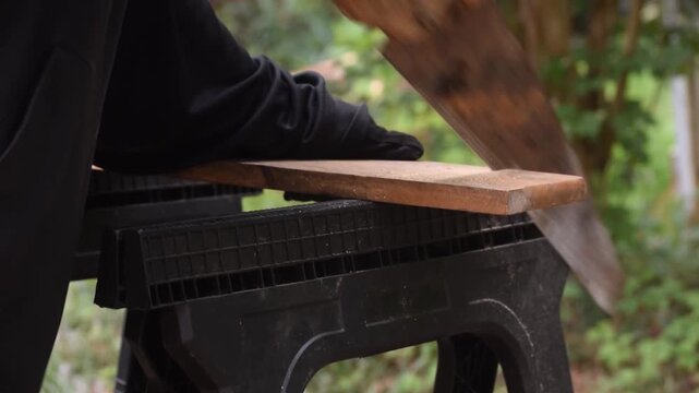 Close-up angled view of a person using an old hand saw to cut a wooden board supported on sawhorses, showing detailed manual woodworking and carpentry work.
