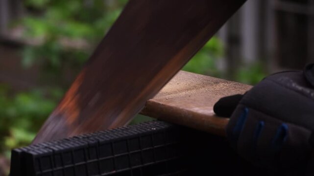 Close-up angled view of a person using an old hand saw to cut a wooden board supported on sawhorses, showing detailed manual woodworking and carpentry work.