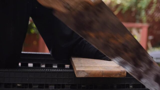 Close-up side view of a person using an old hand saw to cut a wooden board supported on sawhorses, showing detailed manual woodworking and carpentry work.