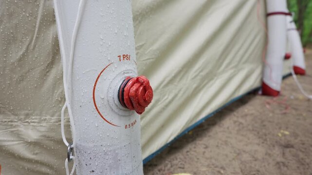 A close-up view shows a red air valve on an inflatable tent beam covered in raindrops. Orange markings indicate the recommended maximum pressure of 7 PSI or 0.5 BAR for safe setup.
