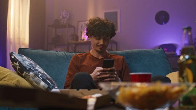 Bearded guy is sitting on sofa. Curly young man starred at screen of smartphone. Guy is concentrated and holding gadget. Boy is wearing smartwatch. Dependence of youth on internet devices.