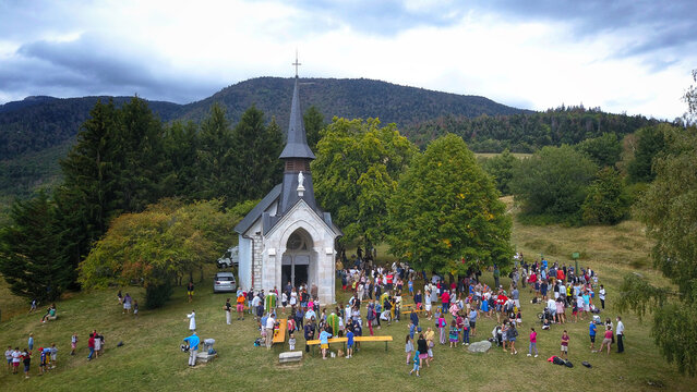 Aerial view of an open-air mass at Chapelle Notre-Dame de Riantmont with people gathered on a grassy hill surrounded by trees and mountains in Vesancy, Auvergne-Rhone-Alpes, France.