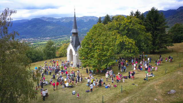 Aerial view of an outdoor mass at Chapelle Notre-Dame de Riantmont with people gathered on a grassy hillside near trees and mountains in Vesancy, Auvergne-Rhone-Alpes, France.