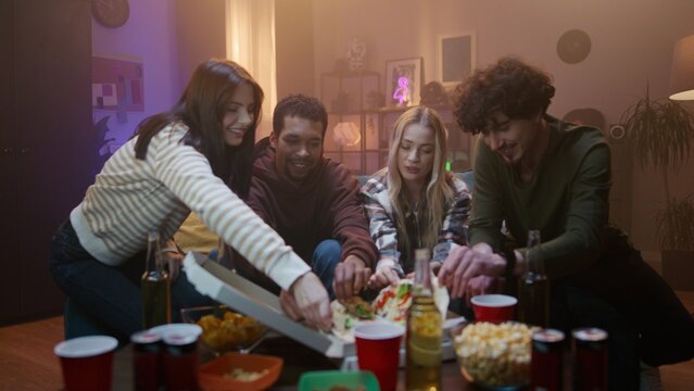 Company of young people is sitting on the sofa. Girl brought pizza for friends. Youth are having fun and enjoying communication. People smile and clap hands. African American man takes slice of pizza.