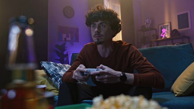 Guy is sitting on sofa and playing video game. Curly-haired young man hold a joystick and gesturing. Male chewing on background of interior and snacks. Evening leisurr of young people.