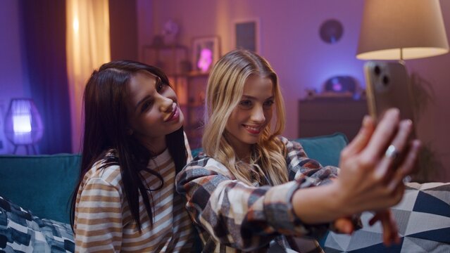 Two joyfull young girls are posing to smartphone. Friends are taking selfie and smiling. Younf women are making post for social network. Best friends sitting on couch and having fun.